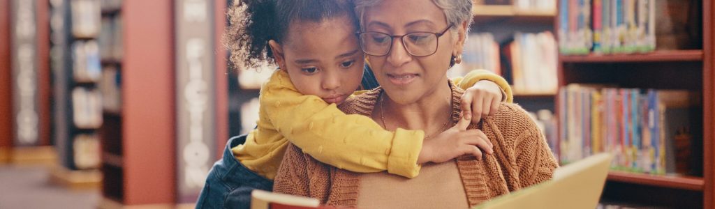 Reading, education and grandma with grandchild in library, hug and bond while sitting together. Learning, storytelling and grandma teaching girl, relax and enjoy books, story and child development